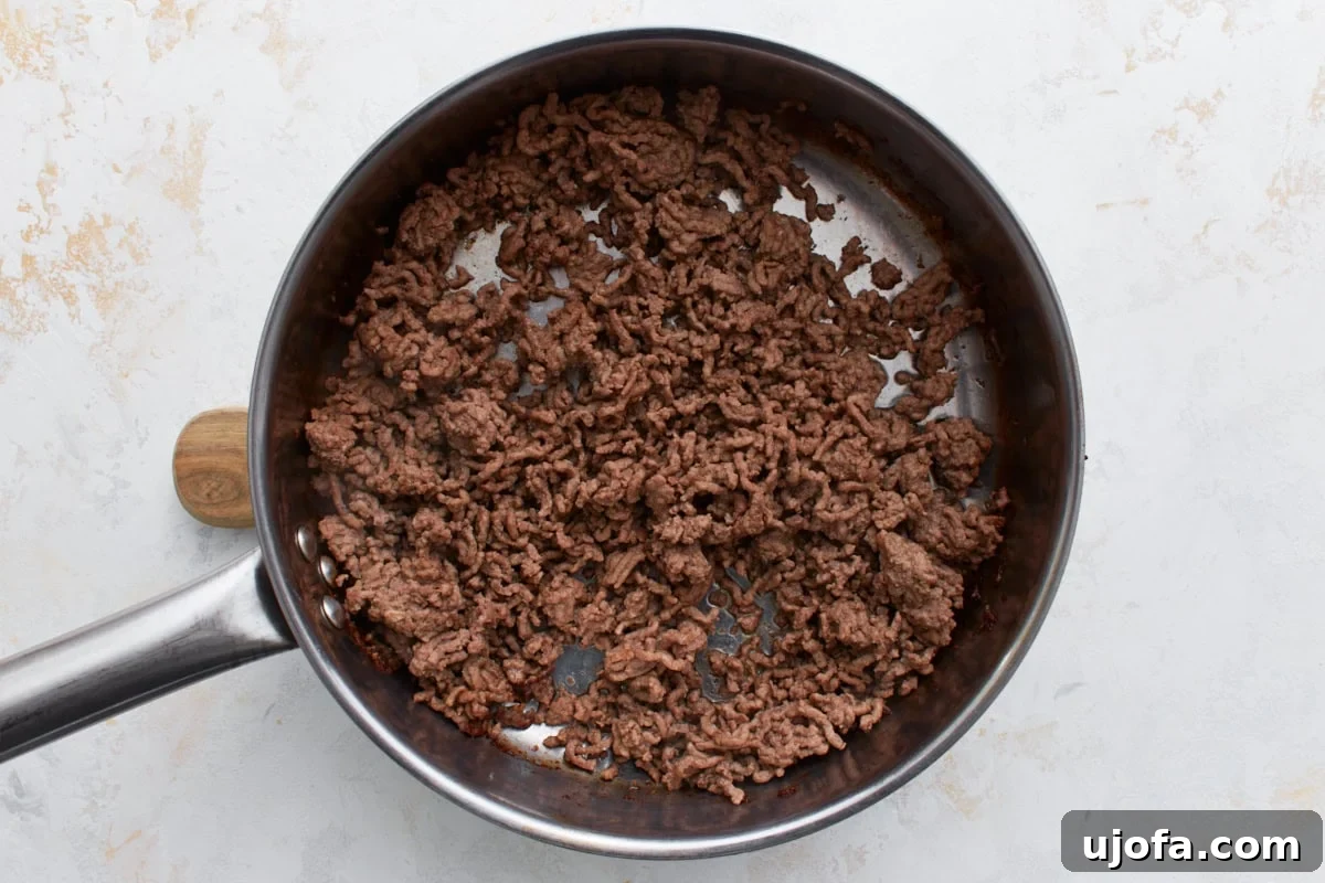 Ground beef fully browned and crumbled in a stainless steel skillet with a wooden handle, resting on a light countertop, indicating it's ready for the next step.