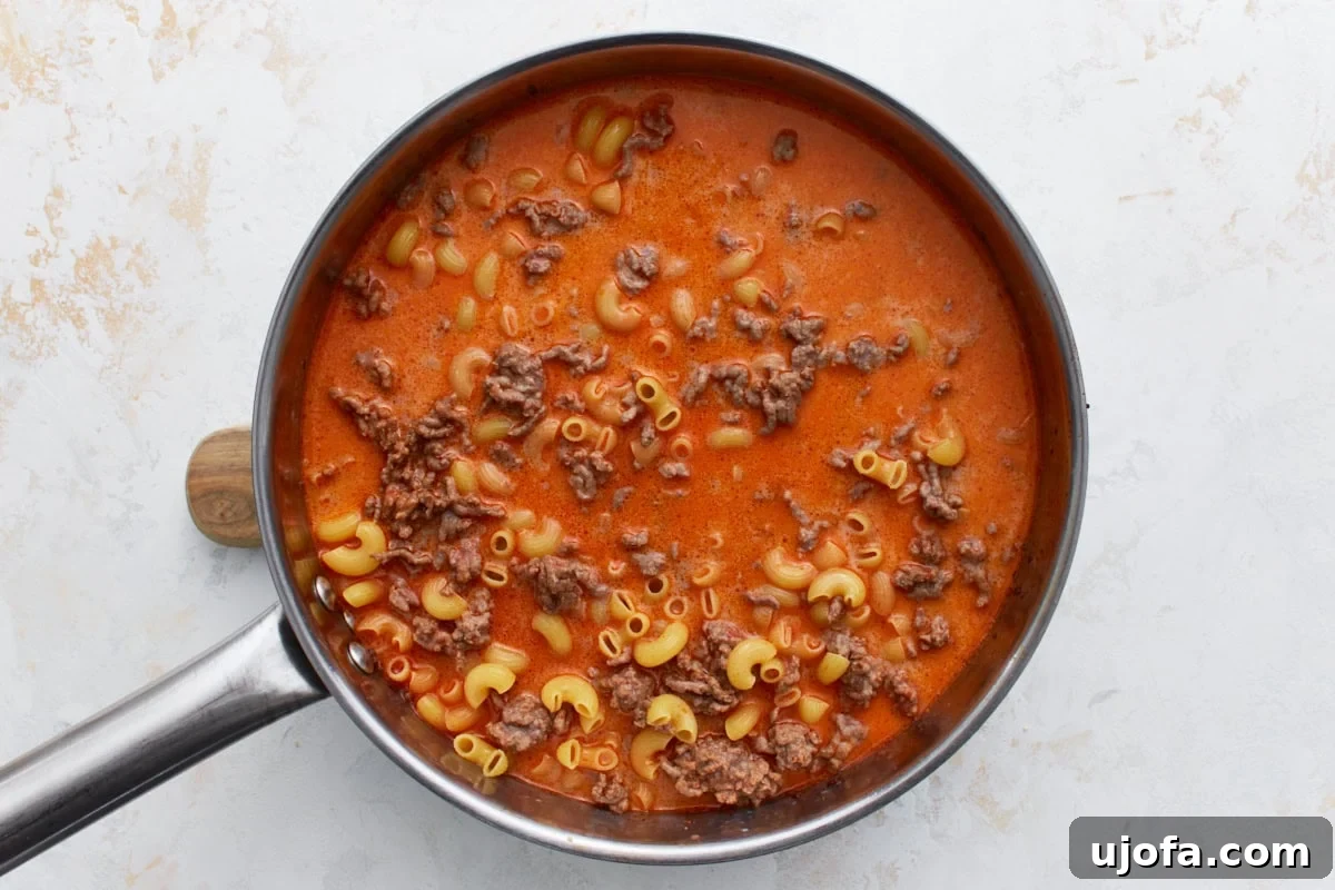 Uncooked gluten-free elbow macaroni pasta submerged in a rich, tomato-based broth with browned ground beef, simmering gently in a large skillet.