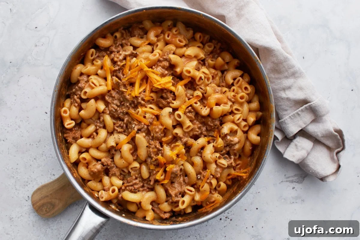 Creamy gluten-free Hamburger Helper, fully cooked and richly sauced, topped with a generous layer of shredded cheddar cheese and a sprinkle of black pepper, still in the skillet.