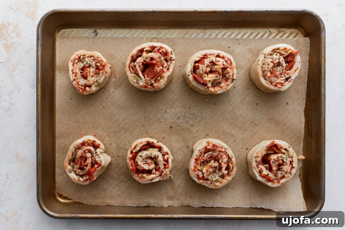 Eight uncooked gluten-free pizza rolls arranged on a parchment-lined baking sheet, with olive oil and oregano brushed on top, ready for the oven.