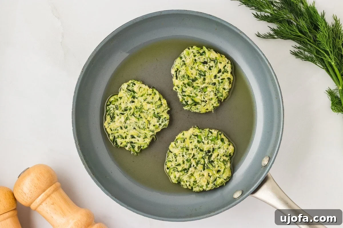 Three raw zucchini fritter patties placed in a skillet with hot oil, frying on their first side.