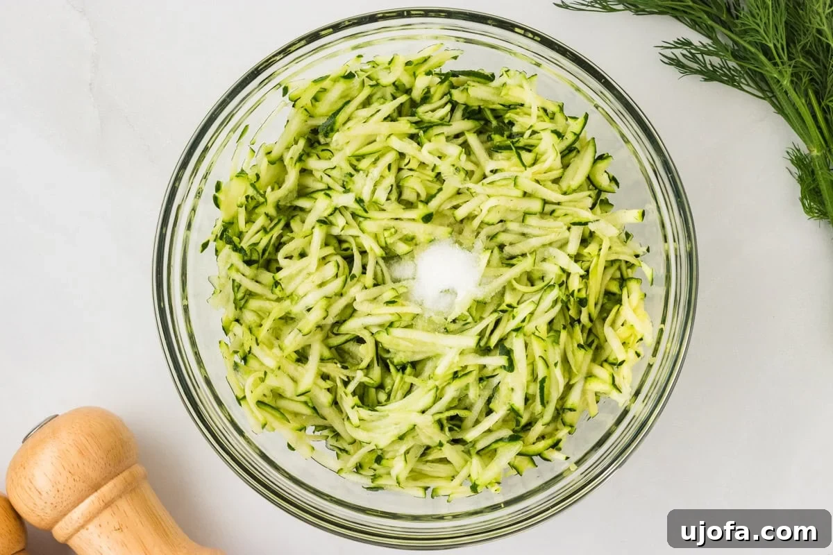A glass bowl filled with freshly grated zucchini sprinkled with salt, ready for the moisture-drawing step.