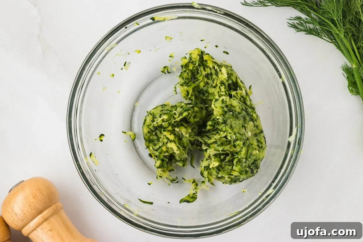 Drained and squeezed zucchini in a glass bowl, formed into a dense, slightly clumped pile after moisture removal.