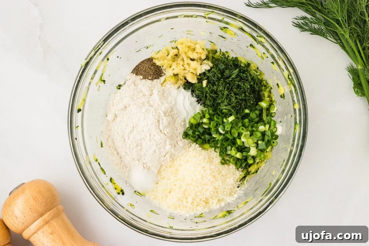 All fritter ingredients—gluten-free flour, parmesan, garlic, dill, green onions, pepper, and salt—layered in a glass bowl with the zucchini.
