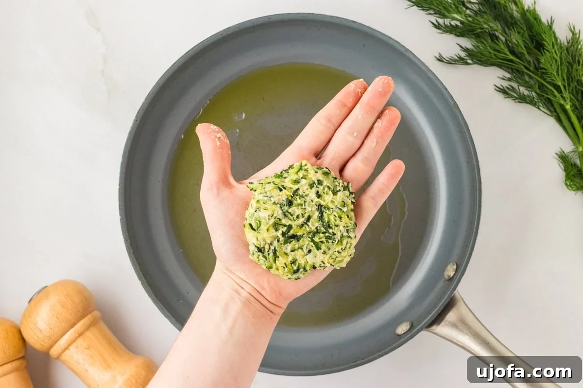 A hand holding a shaped zucchini fritter patty above a skillet coated with oil, ready for frying.