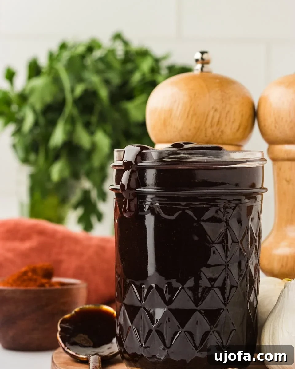 Handcrafted Gluten-Free Barbecue Zest 2 A glass jar filled with thick, dark homemade BBQ sauce, with sauce dripping slightly over the rim. Fresh parsley, garlic, and wooden pepper mills are in the background.