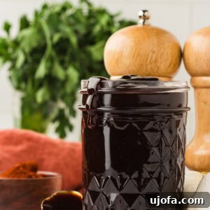 A glass jar filled with thick, dark homemade BBQ sauce, with sauce dripping slightly over the rim. Fresh parsley, garlic, and wooden pepper mills are in the background.