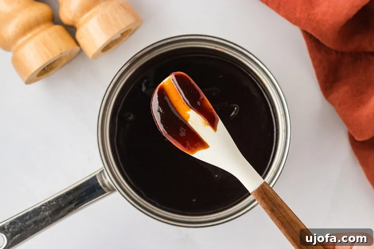 Handcrafted Gluten-Free Barbecue Zest 7 A close-up of a white spoon with wooden handle coated in a glossy stripe of thick BBQ sauce, held above a pot of the finished sauce.