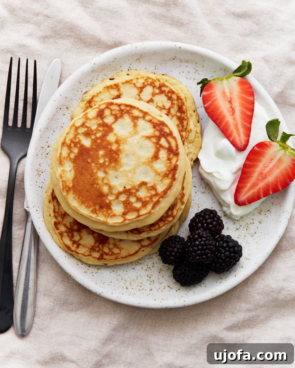 A stack of golden gluten-free protein pancakes on a white plate, served with blackberries, halved strawberries, and a dollop of Greek yogurt. A fork and knife sit beside the plate, inviting you to indulge in this healthy and delicious breakfast.