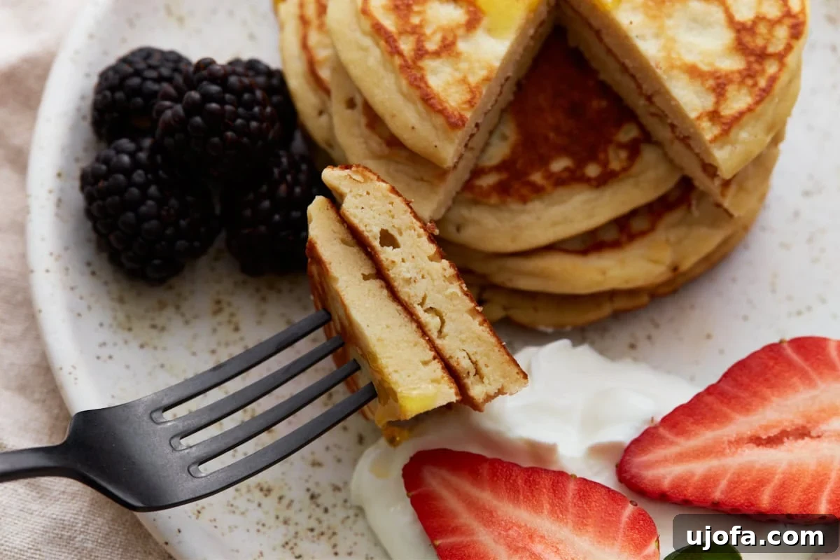 A fork holding a perfect bite of fluffy pancake, layered with Greek yogurt and a strawberry, poised in front of a stack of pancakes, berries, and a knife, showcasing the delightful texture and fresh toppings.