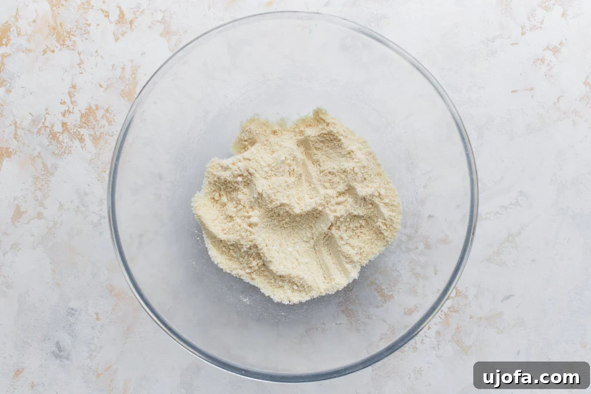 A glass mixing bowl filled with almond flour on a light, textured surface, ready for mixing with other dry ingredients.