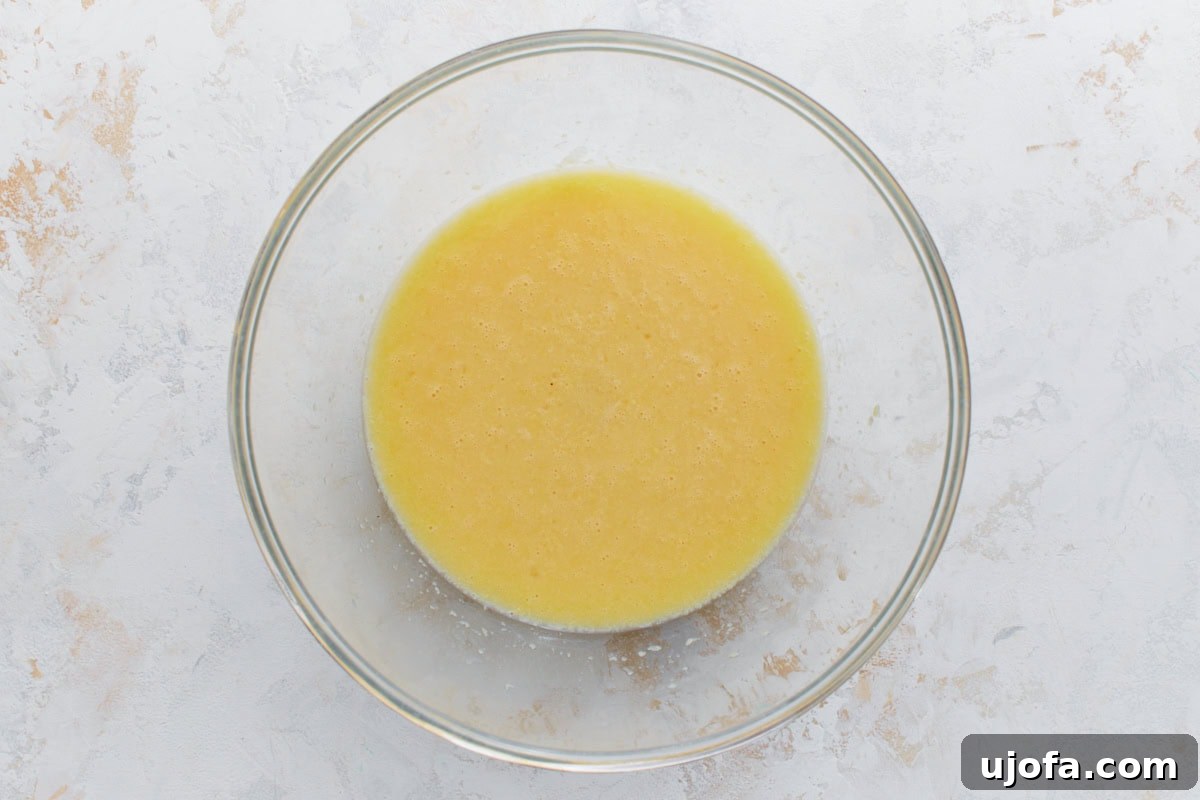 A glass bowl containing a smooth, creamy mixture of wet ingredients like eggs, almond milk, melted butter, maple syrup, and vanilla extract.