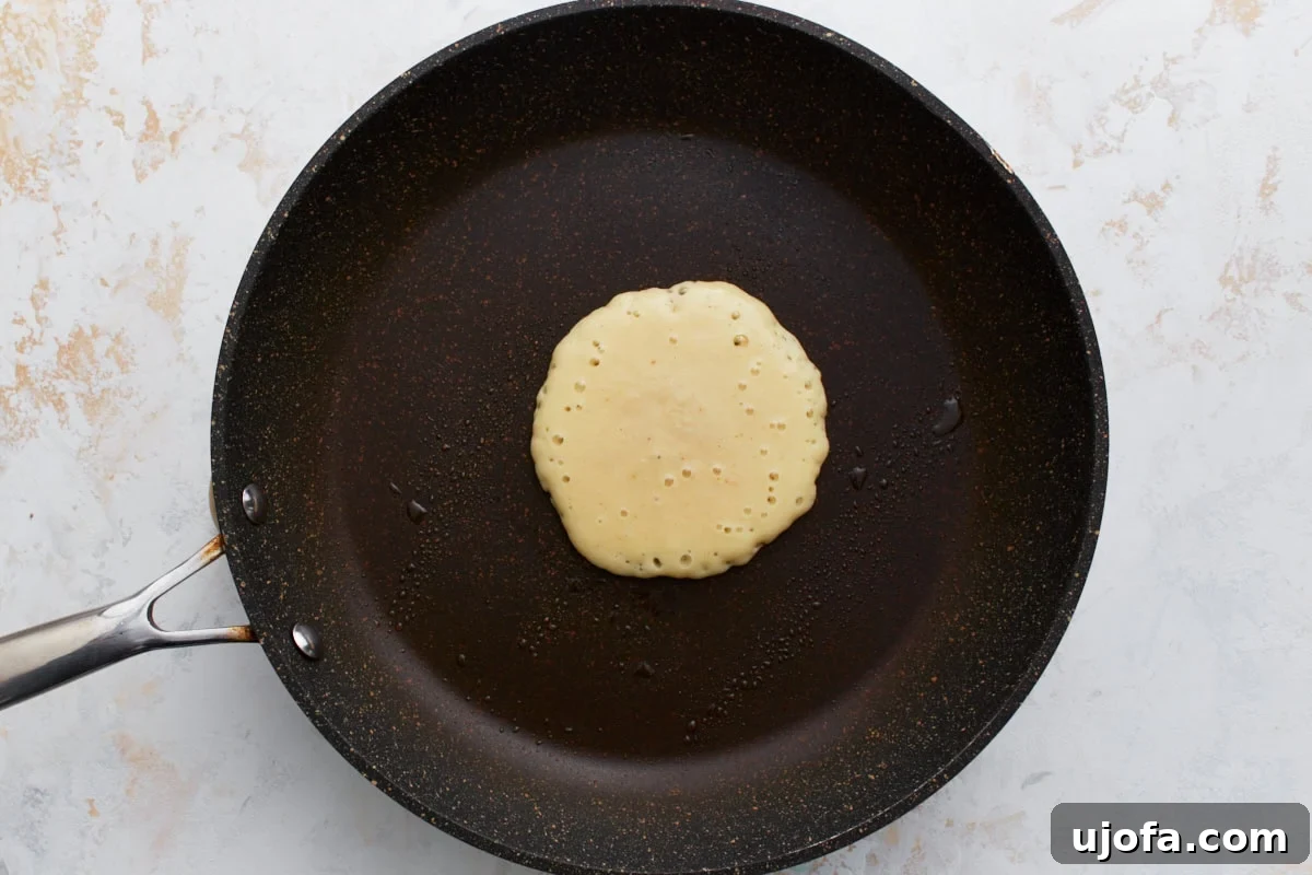 A single pancake cooking in a non-stick skillet, with small bubbles just beginning to form on its golden-brown surface, signaling it's almost ready to flip.