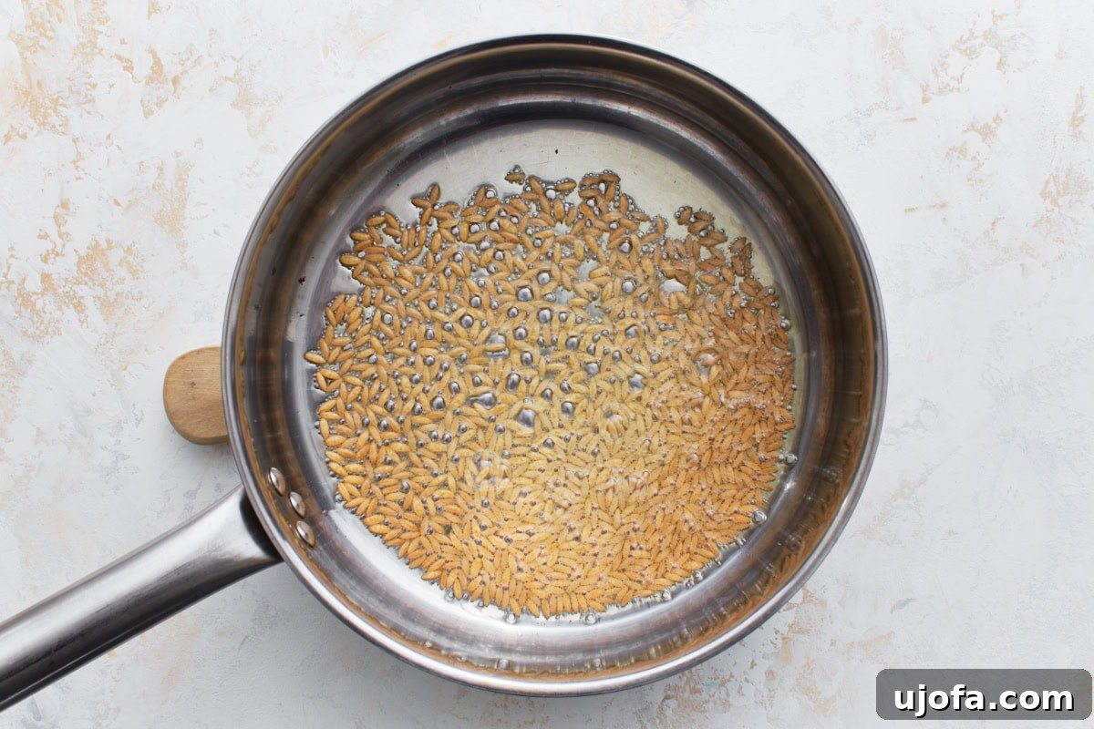 Gluten-free orzo pasta being toasted to a golden brown in olive oil within a stainless steel pan, stirred for even cooking.
