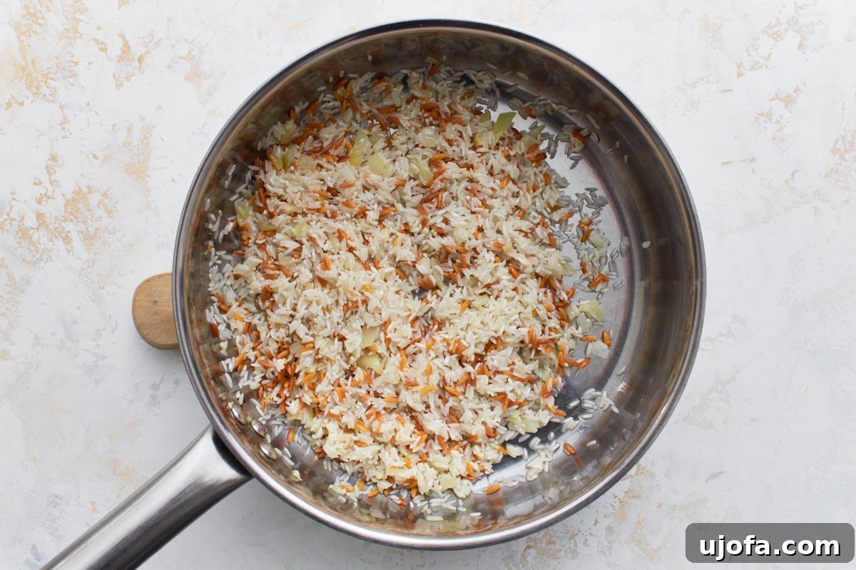 Rinsed long-grain rice being stirred into the pan with toasted orzo and sautéed onions and garlic, combining all elements before liquid is added.