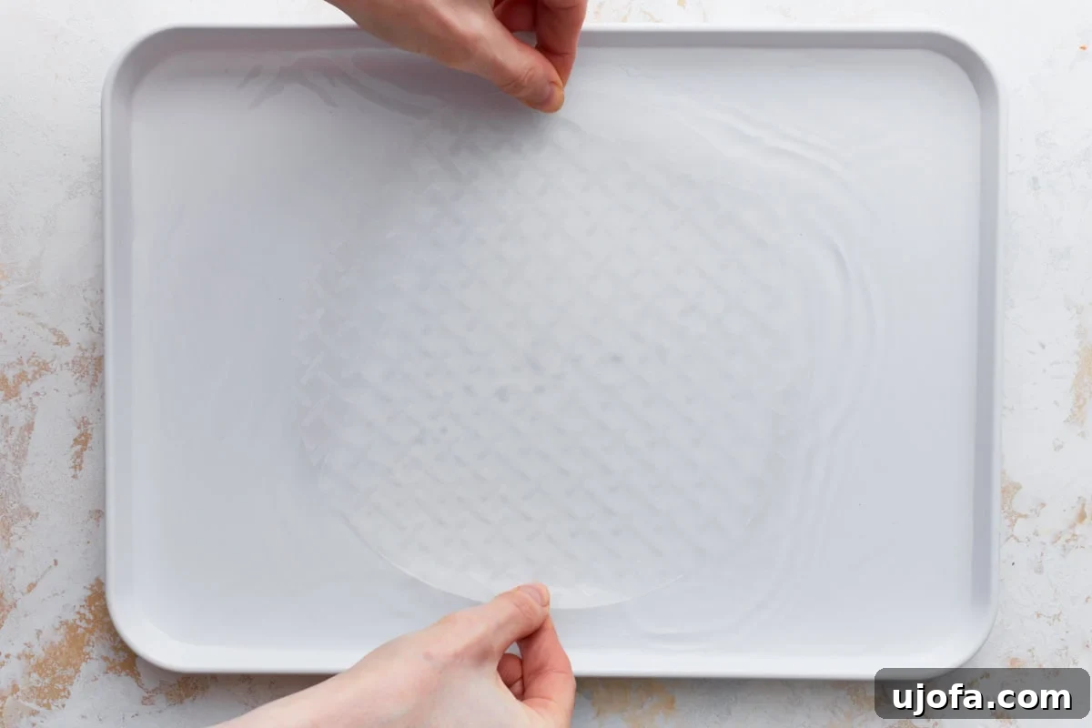 A person dips a round sheet of rice paper into a tray of warm water to soften it for rolling.