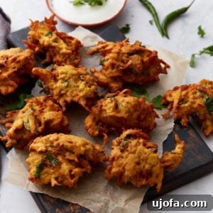 Golden-brown pakoras arranged on parchment paper over a dark wooden board, served with a bowl of creamy white dipping sauce and fresh herbs.