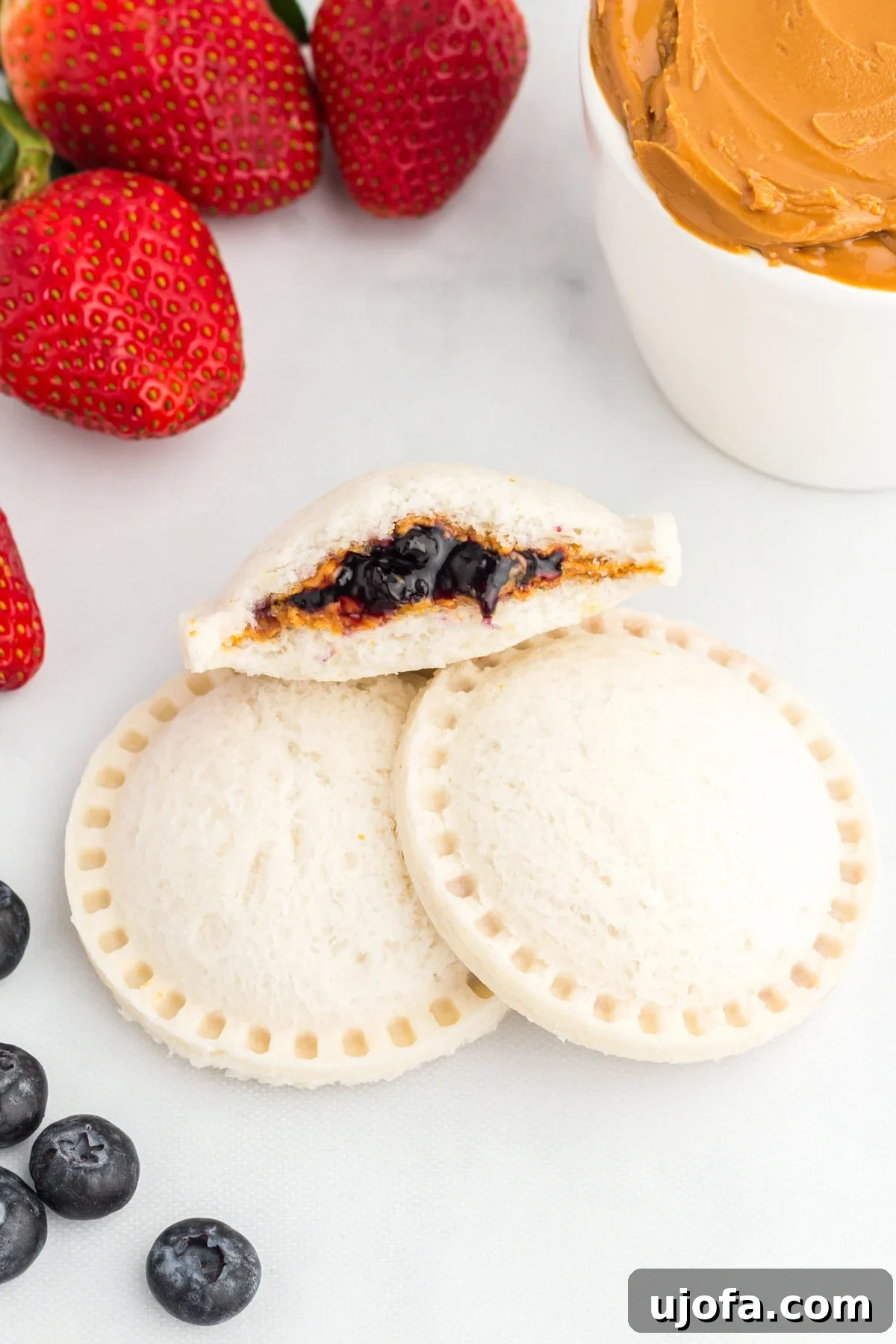 GF Lunchbox Wonders 2 Two gluten-free uncrustables stacked with one cut open to show the peanut butter and blueberry jam filling, surrounded by fresh strawberries, blueberries, and a small bowl of peanut butter.