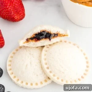 Two gluten-free uncrustables stacked with one cut open to show the peanut butter and blueberry jam filling, surrounded by fresh strawberries, blueberries, and a small bowl of peanut butter.