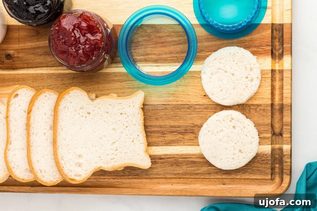 GF Lunchbox Wonders 5 Slices of gluten-free bread laid out on a wooden cutting board, with a blue sandwich cutter, jars of jam, and two bread circles cut out and ready to fill.