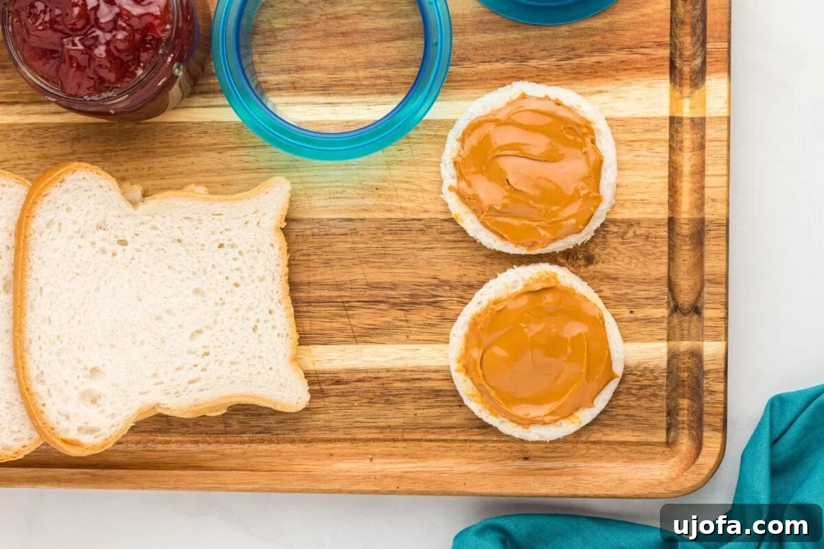 GF Lunchbox Wonders 6 Two gluten-free bread circles spread with a layer of peanut butter on a wooden cutting board, next to more bread slices and jars of jam.