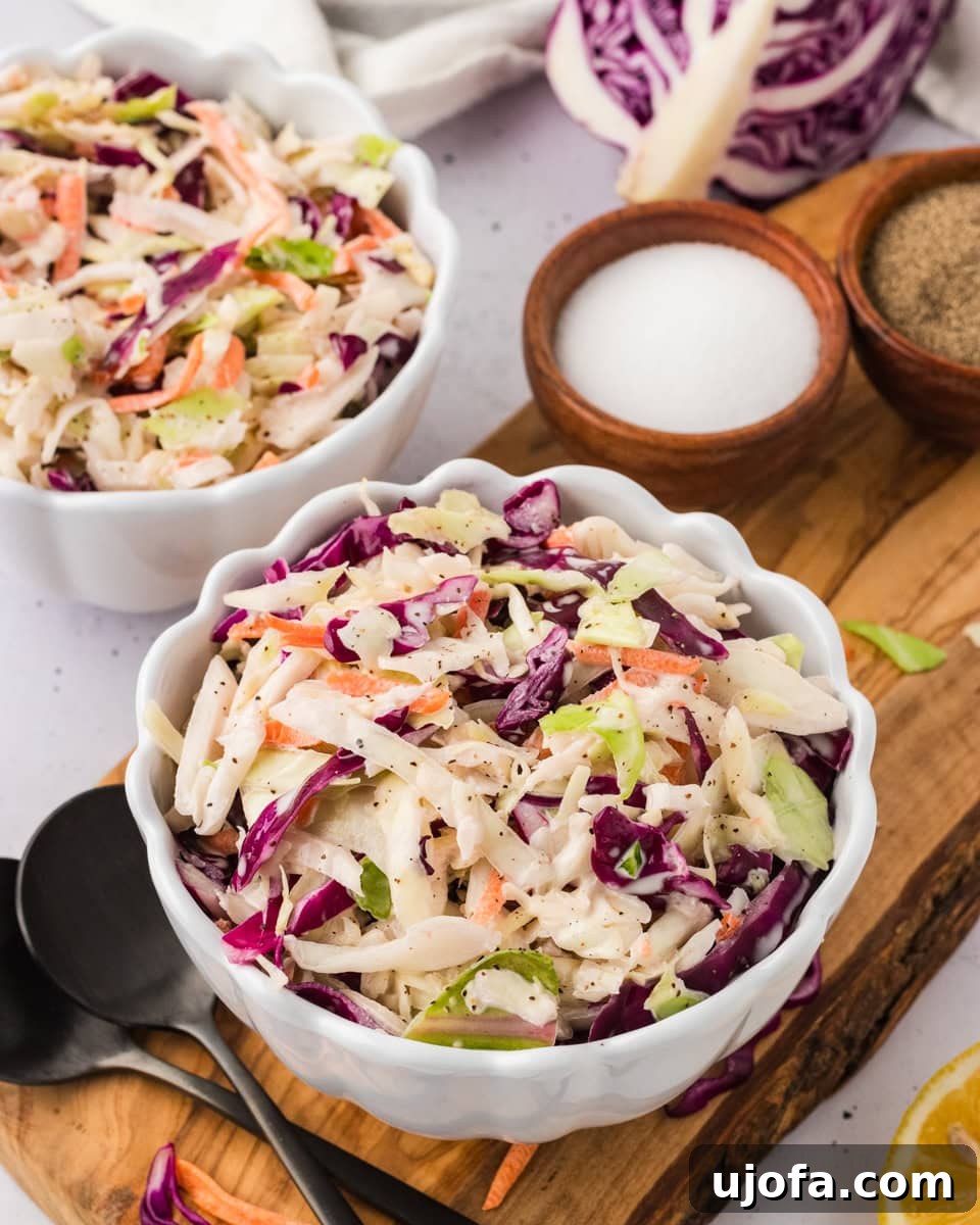 A close-up of two white scalloped bowls filled with colorful dairy-free coleslaw, placed on a wooden board with small bowls of salt and pepper and a halved red cabbage in the background.