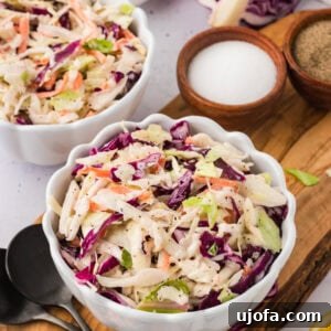 A close-up of two white scalloped bowls filled with colorful dairy-free coleslaw, placed on a wooden board with small bowls of salt and pepper and a halved red cabbage in the background.