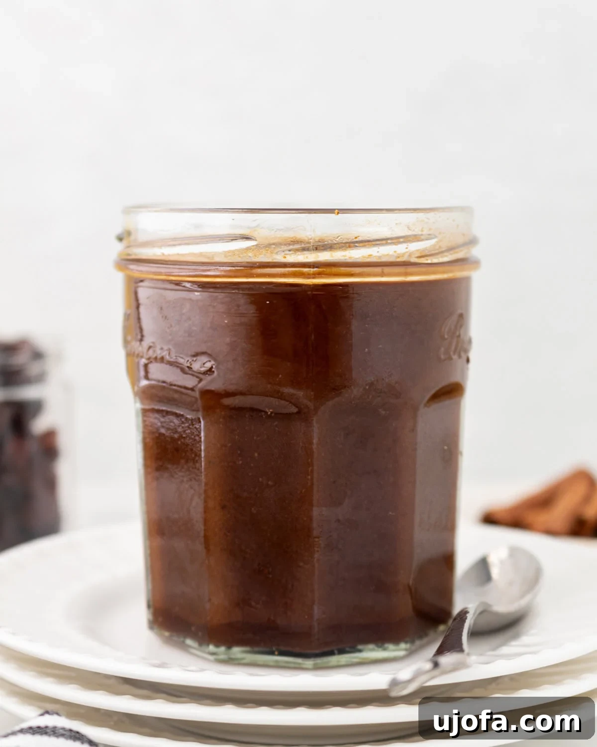 Gluten-Free Worcestershire from Scratch 2 A glass jar filled with dark brown homemade Worcestershire sauce, placed on a stack of white plates with a spoon beside it.