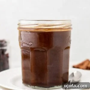 A glass jar filled with dark brown homemade Worcestershire sauce, placed on a stack of white plates with a spoon beside it.