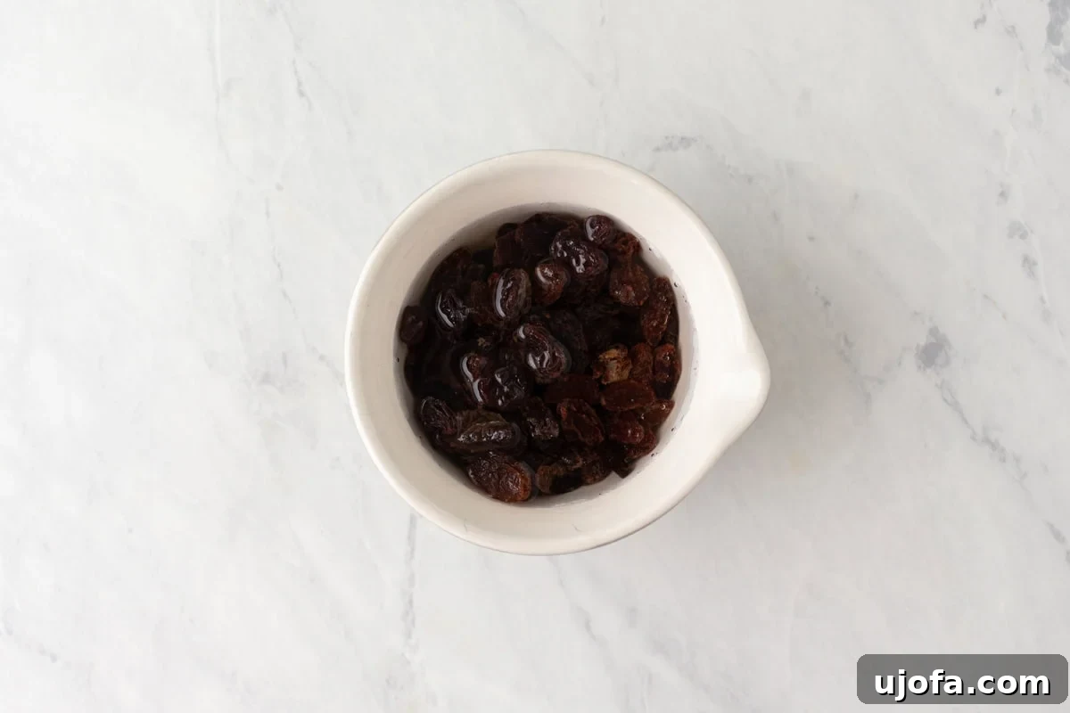 Gluten-Free Worcestershire from Scratch 5 A small white bowl filled with dark raisins soaking in water, sitting on a light marble surface.