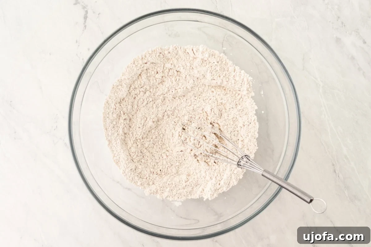 Fluffy Gluten-Free Drop Biscuits 5 A large glass bowl containing a freshly whisked gluten-free flour mixture, with a whisk resting inside, on a pristine white marble surface.