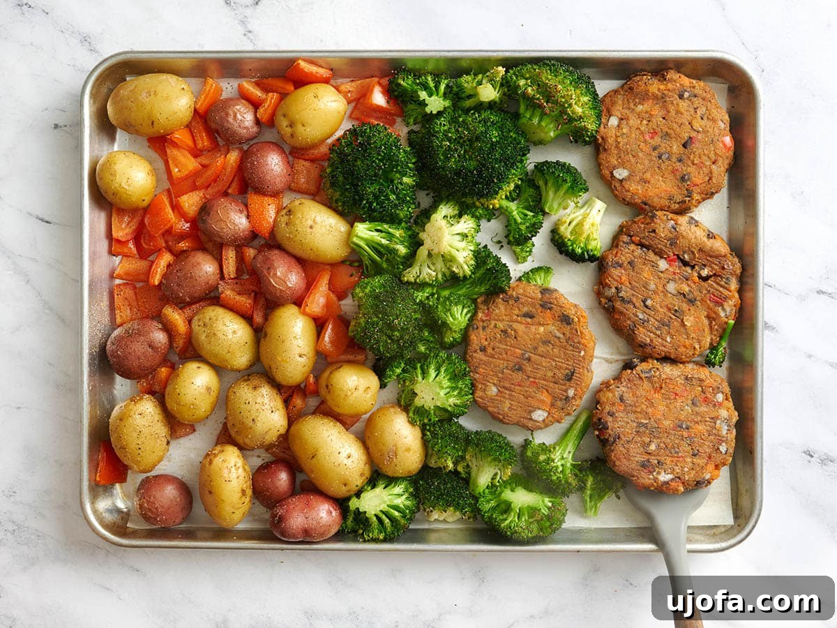 Tangy Tzatziki Black Bean Power Bowl 8 Overhead view of partially cooked potatoes and diced tomatoes, broccoli florets, and black bean burgers on a baking sheet, with a spatula flipping one of the burgers.