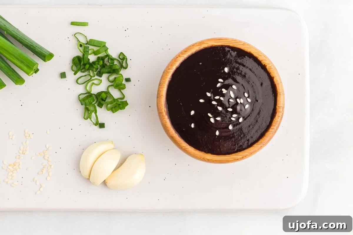 Craft Your Own Gluten-Free Hoisin Sauce 8 Overhead view of a bowl of hoisin sauce topped with sesame seeds, surrounded by fresh green onions, garlic cloves, and sesame seeds on a white background.
