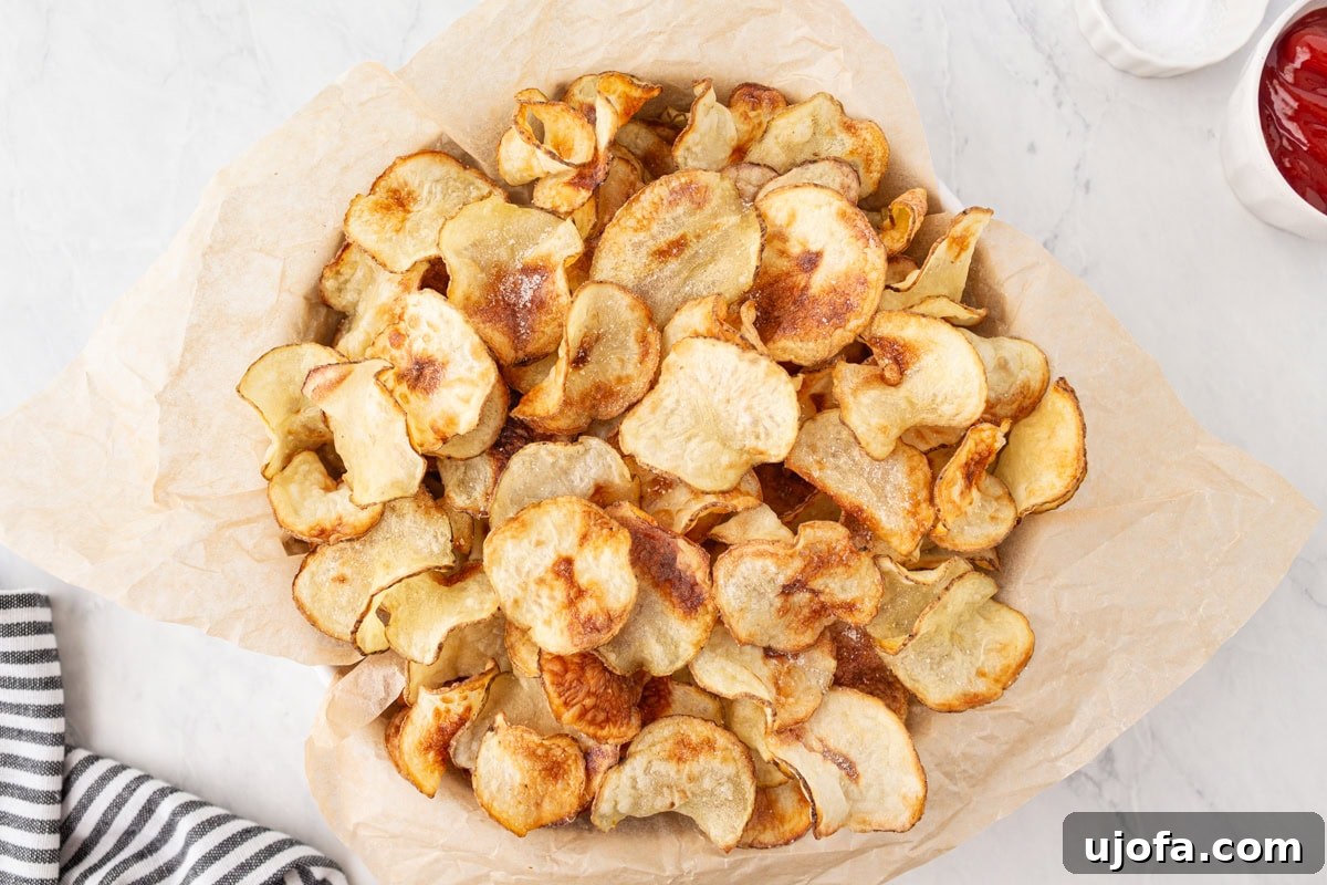 A generous serving of homemade gluten-free potato chips, presented in a parchment-lined bowl, accompanied by a small ramekin of ketchup for dipping.