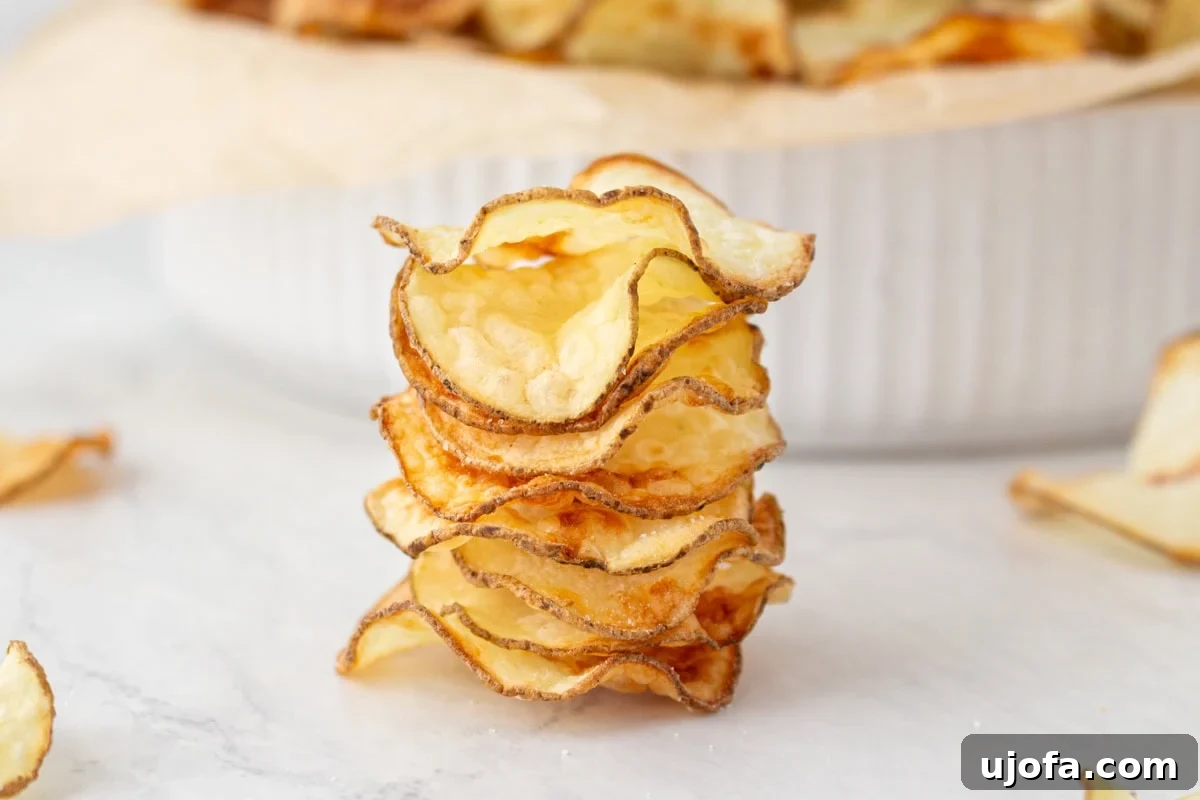 A close-up stack of several homemade crispy potato chips, highlighting their wavy, airy texture and golden color, set against a pristine white marble background.