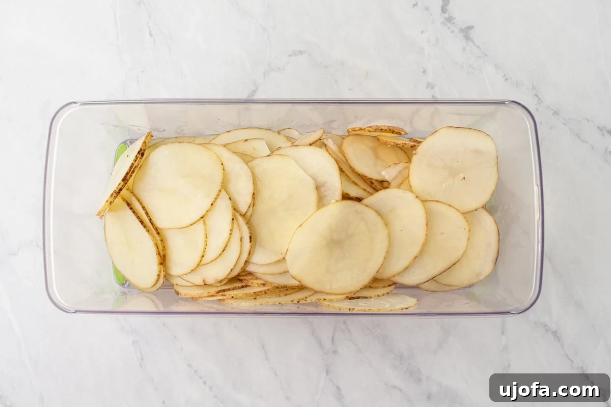 Thinly sliced raw russet potatoes arranged neatly in a clear glass container, showcasing their uniform 1/16-inch thickness after being processed with a mandolin.
