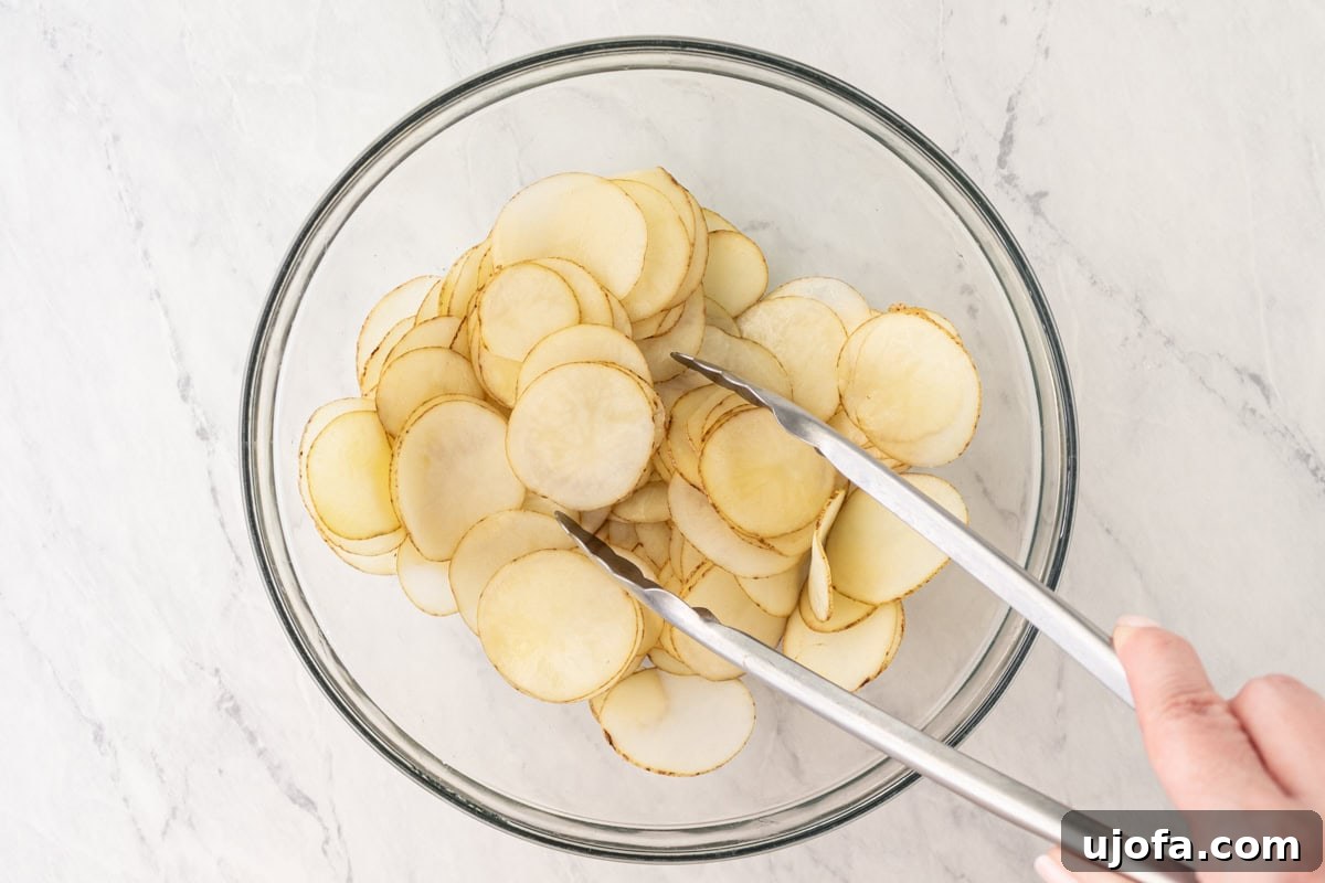 Potato slices in a clear glass bowl being actively tossed with tongs, ensuring an even coating of olive oil and seasoning before air frying.