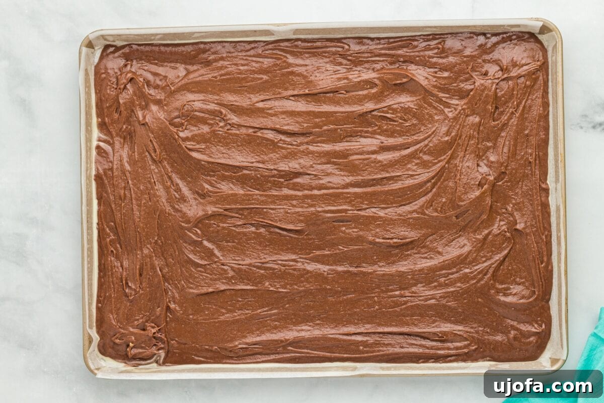 Thick chocolate cookie batter expertly spread into an even layer on a parchment-lined rimmed baking sheet, ready to be baked into the base for ice cream sandwiches.