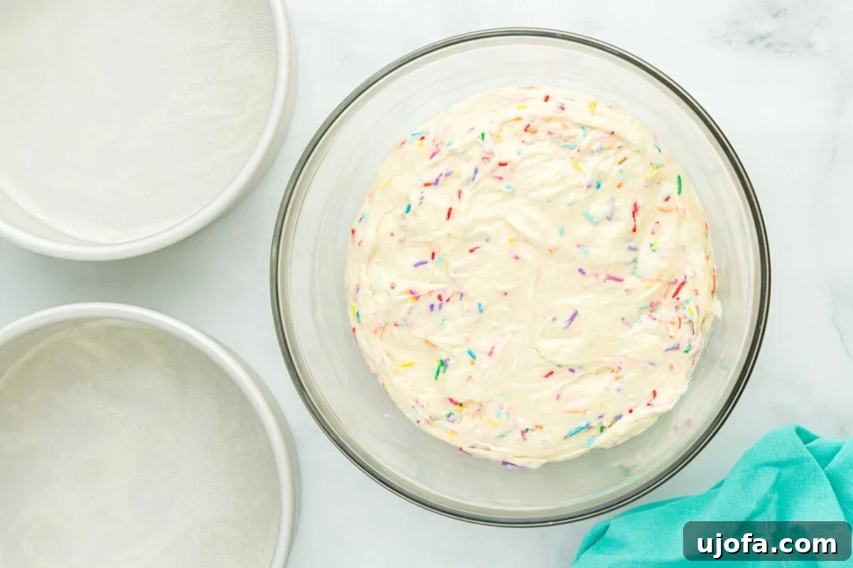 A bowl of thick gluten-free cake batter with colorful sprinkles folded in, next to prepared round cake pans.