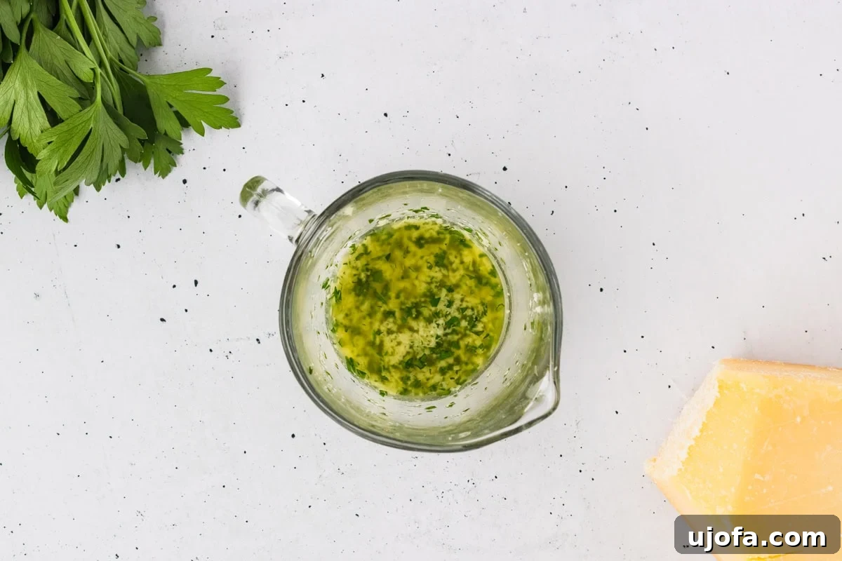 A small glass measuring cup filled with aromatic melted garlic herb butter, accompanied by fresh parsley and a wedge of Parmesan cheese, ingredients for the breadstick topping.