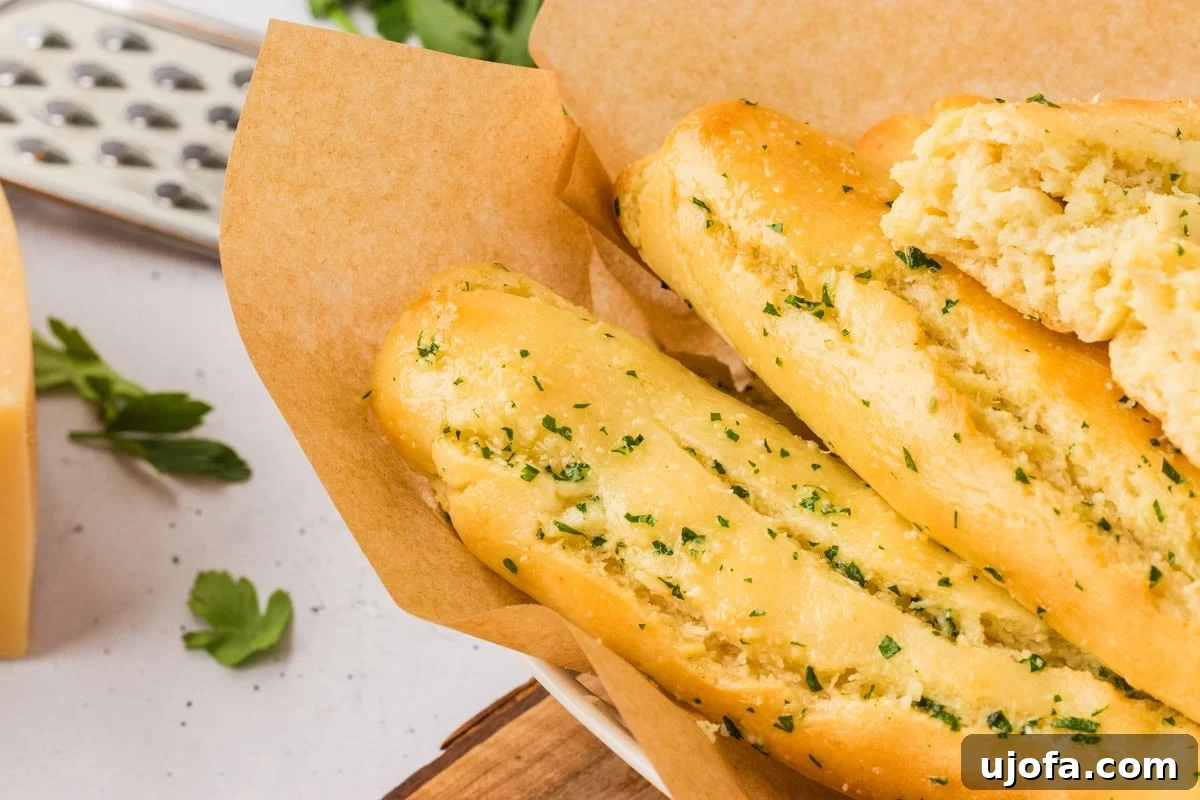 A rustic bowl filled with golden, buttery gluten-free breadsticks, lined with parchment paper. One breadstick is broken open to reveal its perfectly soft, airy texture within.