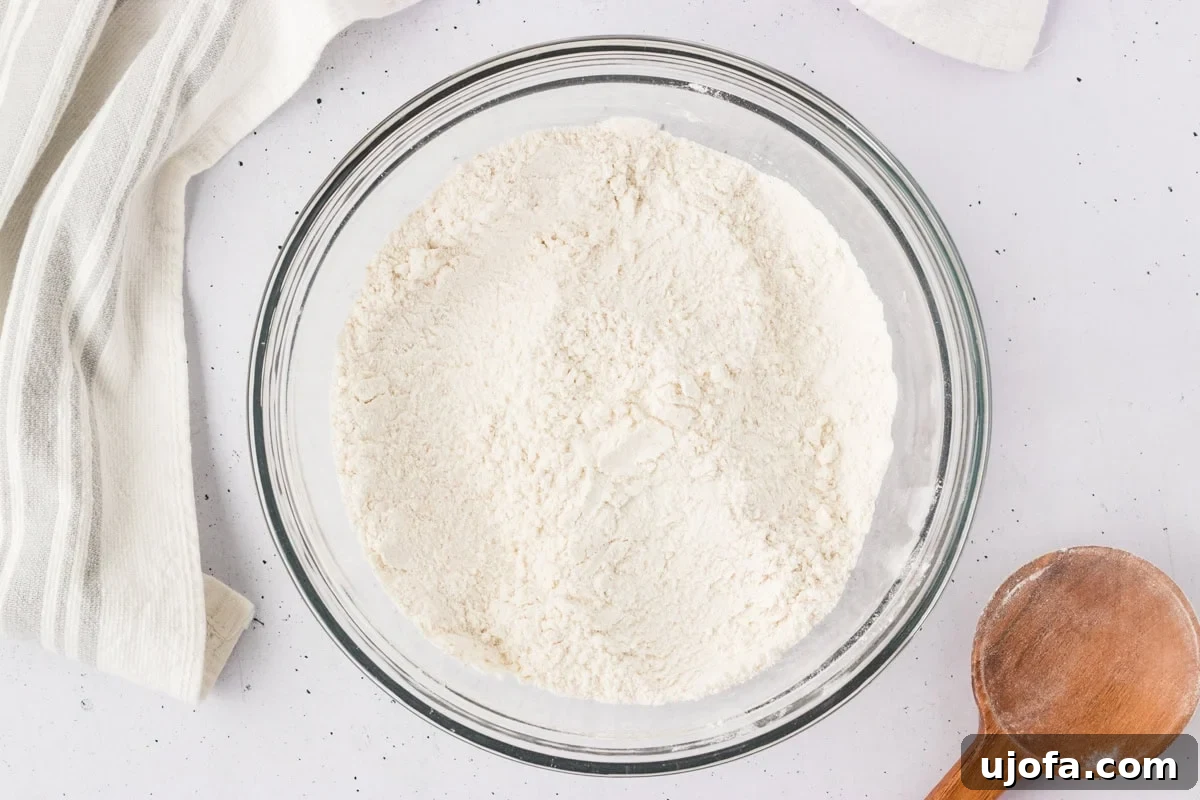 Overhead view of a clear glass bowl filled with a well-combined gluten-free flour mixture, including yeast, sugar, and salt, on a white countertop.