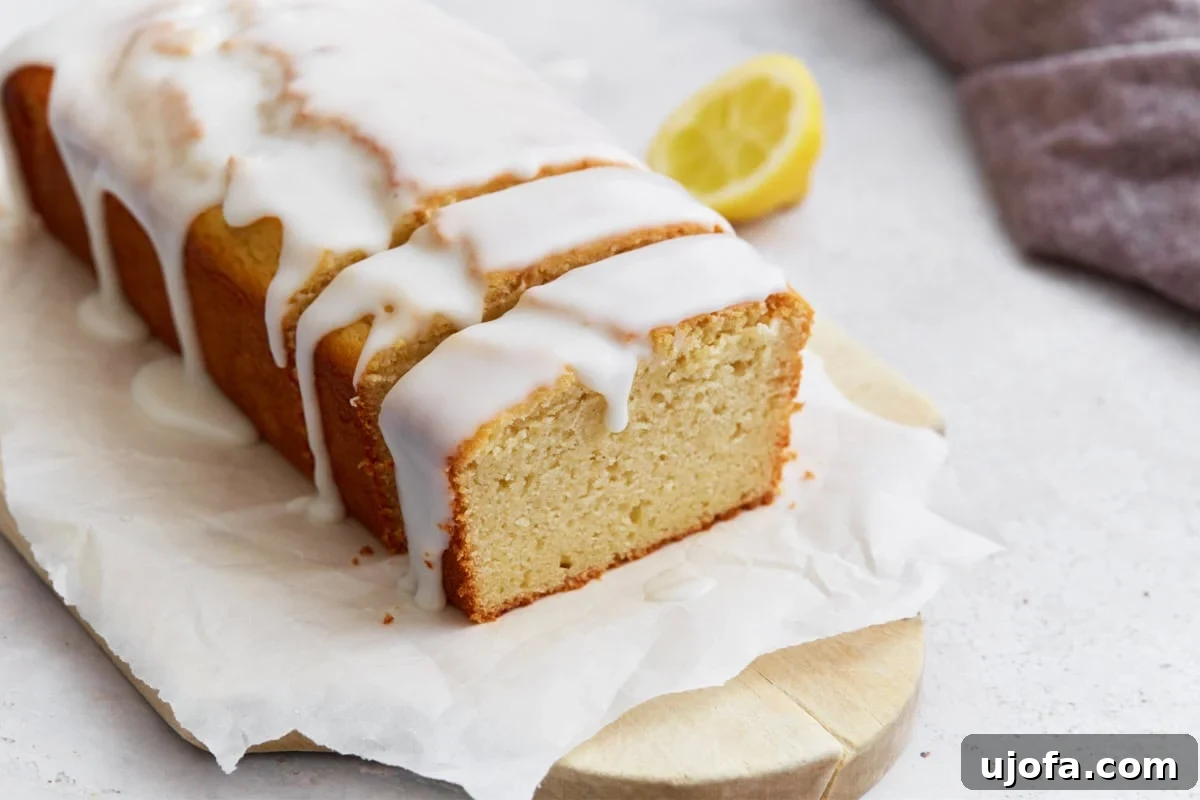 Sliced gluten-free iced lemon loaf on parchment paper, with a lemon half in the background and glaze dripping down the sides.