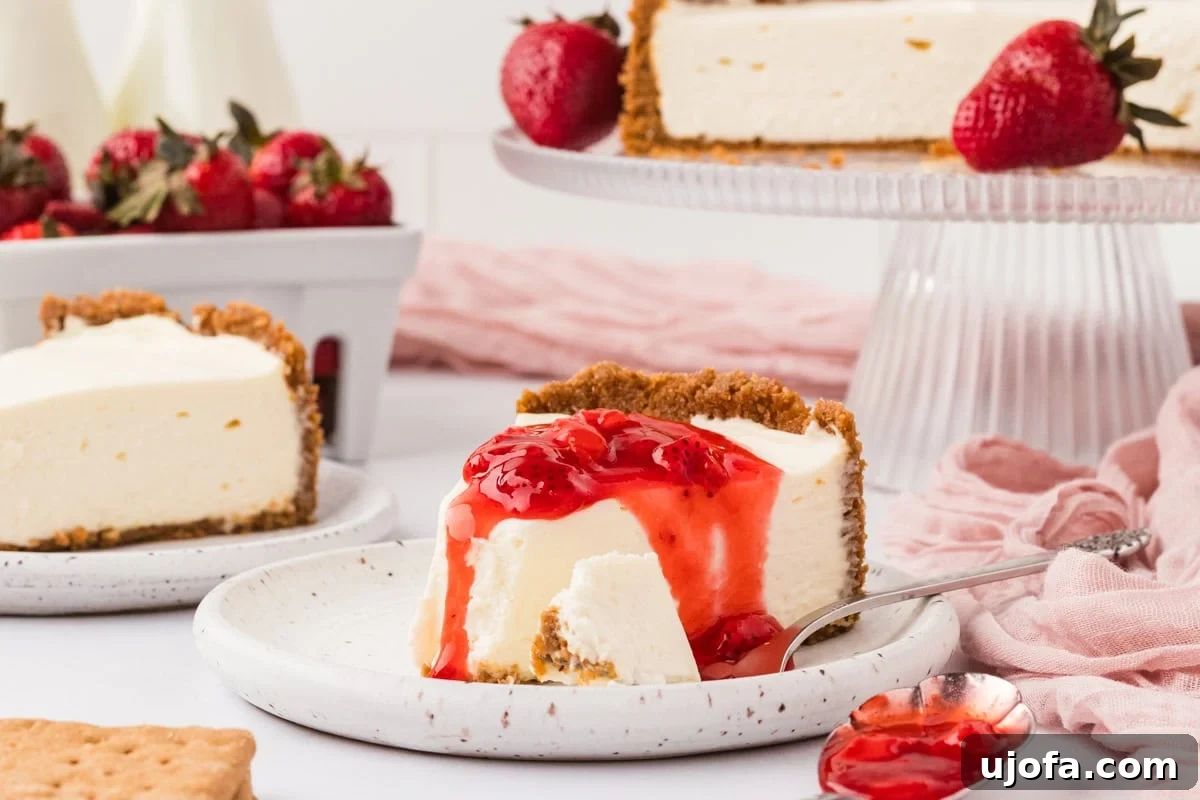 No-Oven Gluten-Free Cheesecake Bliss 14 A slice of gluten-free no-bake cheesecake topped with strawberry sauce, with the whole cheesecake and fresh strawberries in the background on a white and pink styled table.