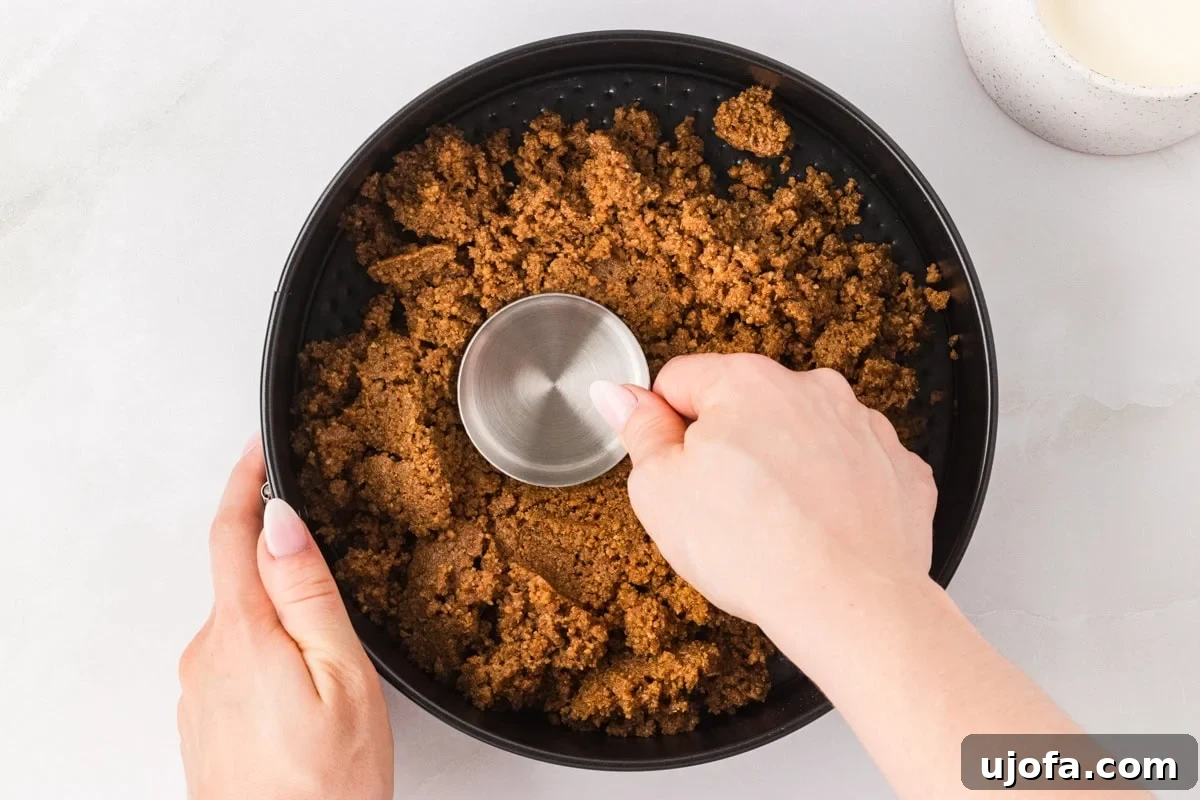 No-Oven Gluten-Free Cheesecake Bliss 6 A pair of hands firmly pressing the graham cracker crust mixture into the bottom and up the sides of a 9-inch springform pan using a sturdy metal measuring cup, ensuring an even and compact base.