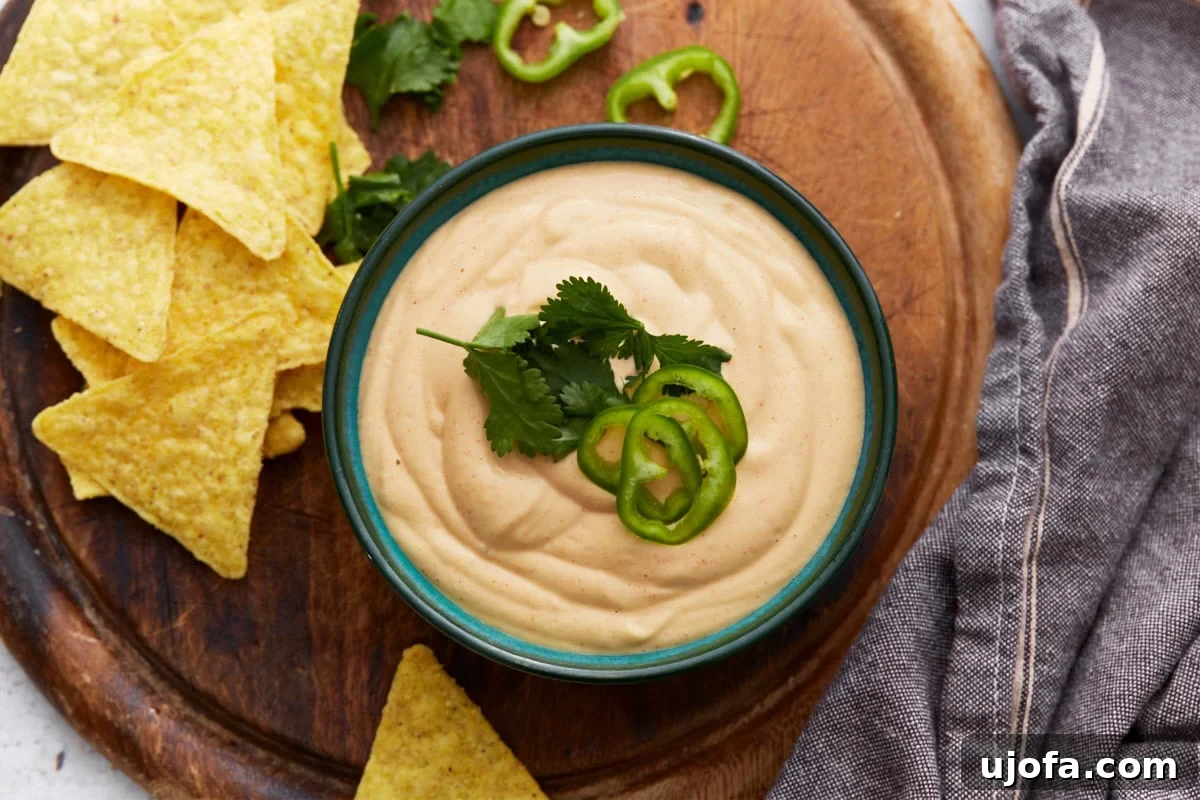 Creamy Cashew Queso 7 A beautifully presented bowl of cashew queso dip, garnished with vibrant cilantro and thin jalapeño slices, accompanied by a pile of golden tortilla chips on a wooden serving board.