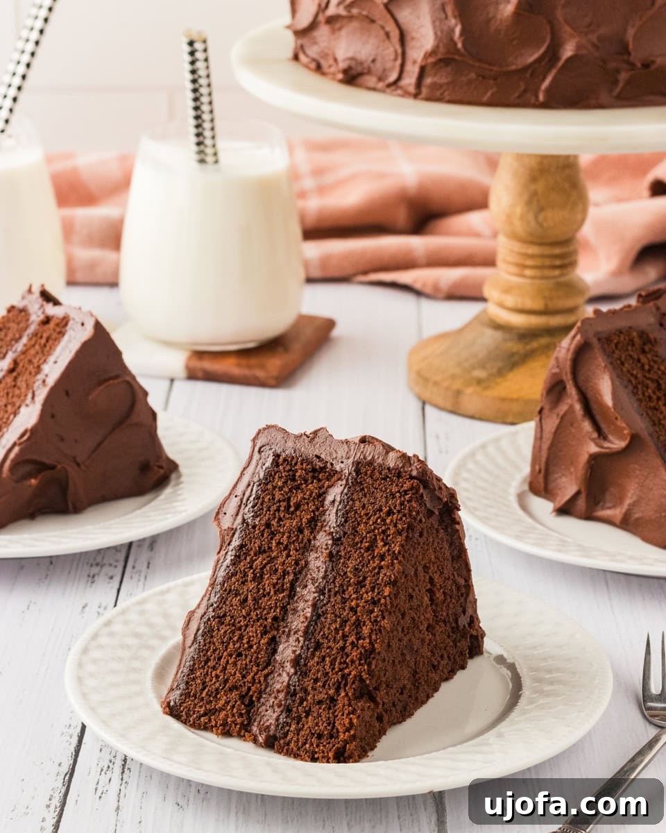 Effortless Gluten-Free Fudgy Cake 2 A slice of rich, two-layer chocolate cake with creamy chocolate frosting, served on a white plate. A full cake sits on a wooden cake stand in the background, with additional slices plated and glasses of milk nearby.