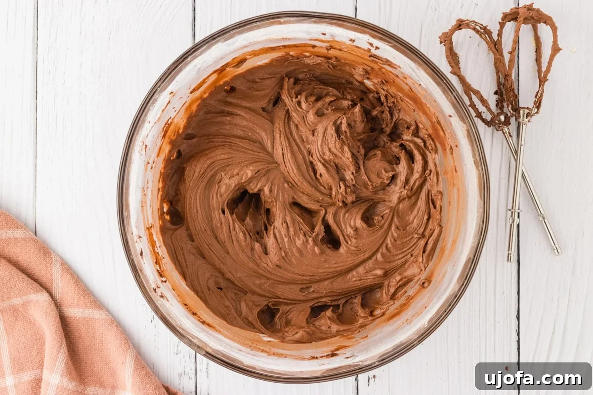 Effortless Gluten-Free Fudgy Cake 18 A glass bowl with fully mixed chocolate frosting, smooth and fluffy, with a hand mixer coated in frosting resting beside it.