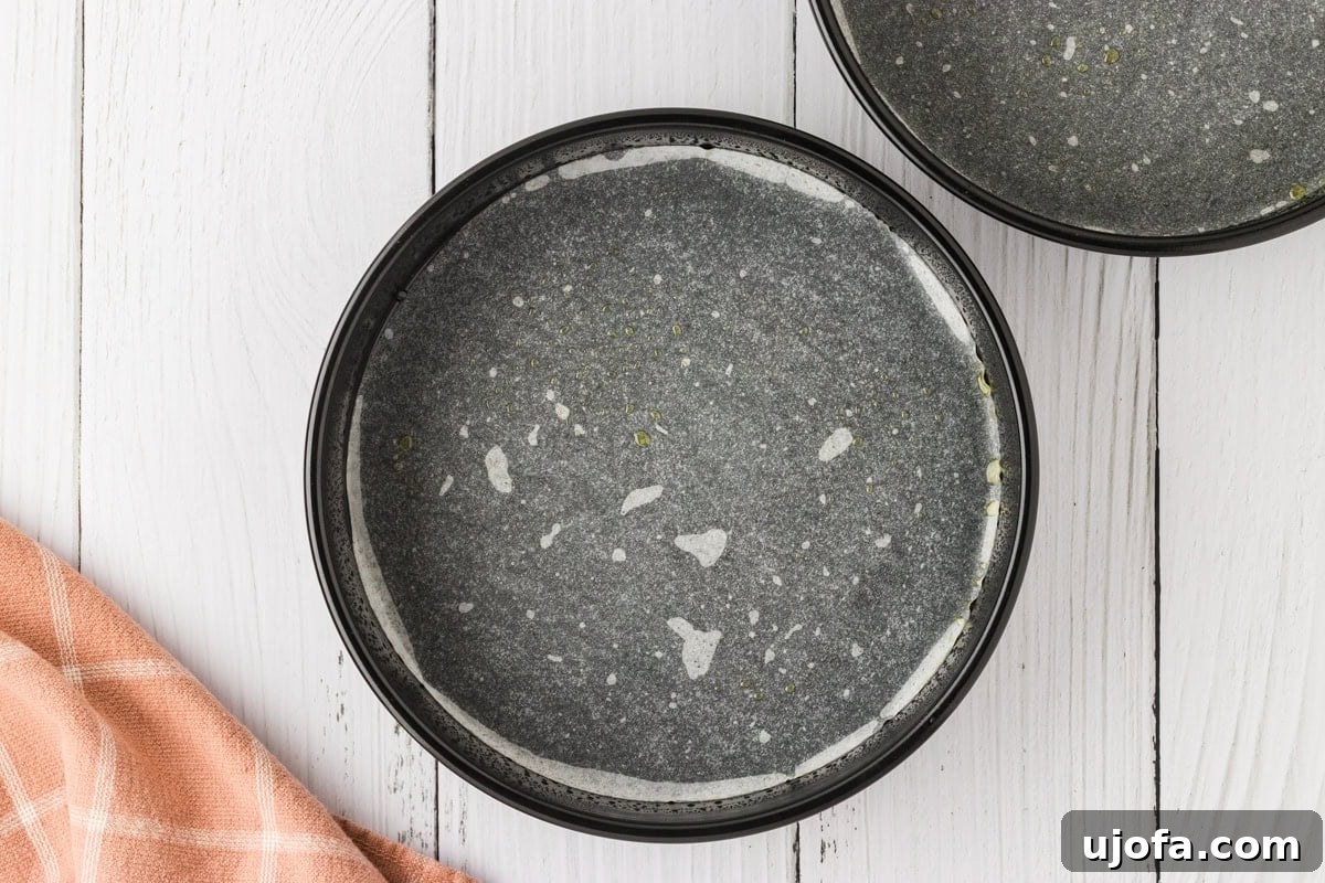 Effortless Gluten-Free Fudgy Cake 5 Two round cake pans lined with parchment paper and lightly greased, sitting on a white wooden surface with a soft pink-checkered kitchen towel.