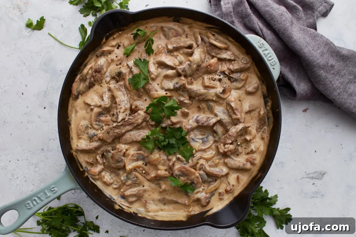 Gluten-free beef stroganoff served in a skillet and garnished with fresh parsley, placed on a white background with scattered herbs and a linen napkin.